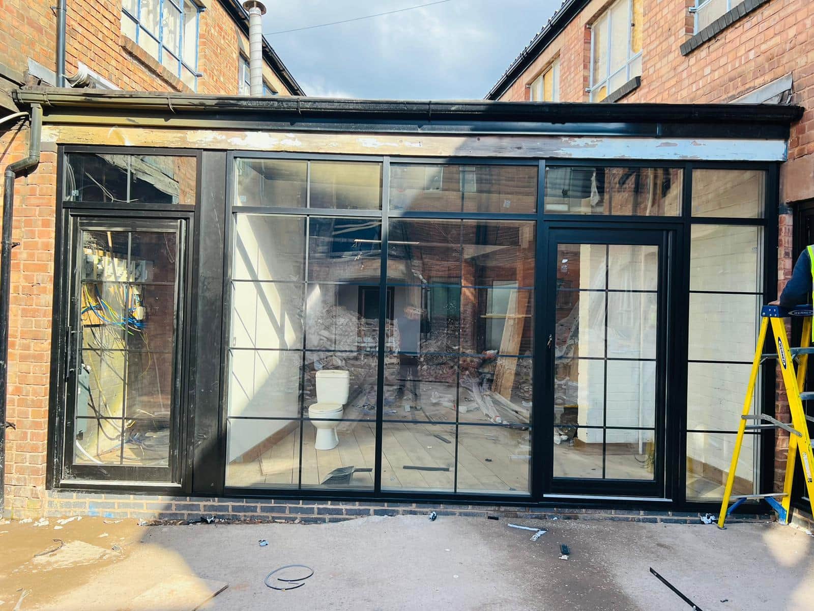 Black-framed glazed screen with doors and slim glazing bars across the rear of a brick building, during installation.