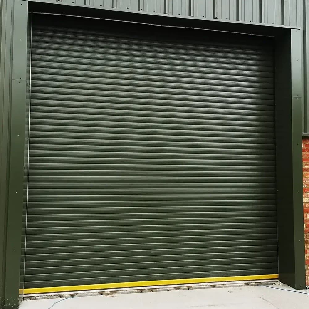 Dark green steel roller shutter closed across a loading opening in a clad industrial unit, with a yellow bottom rail