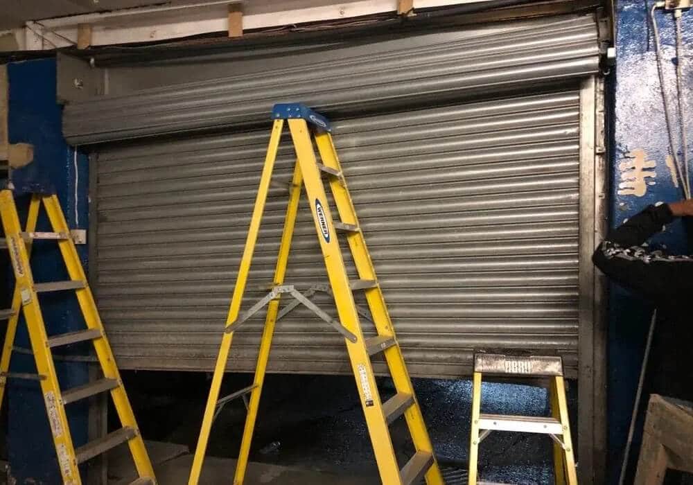 A shop’s roller shutter being worked on, with stepladders set up beneath it.